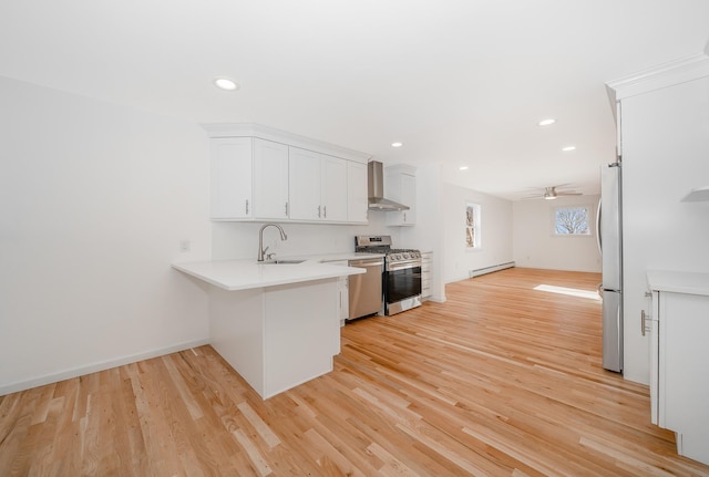 kitchen featuring baseboard heating, appliances with stainless steel finishes, ceiling fan, white cabinetry, and wall chimney range hood