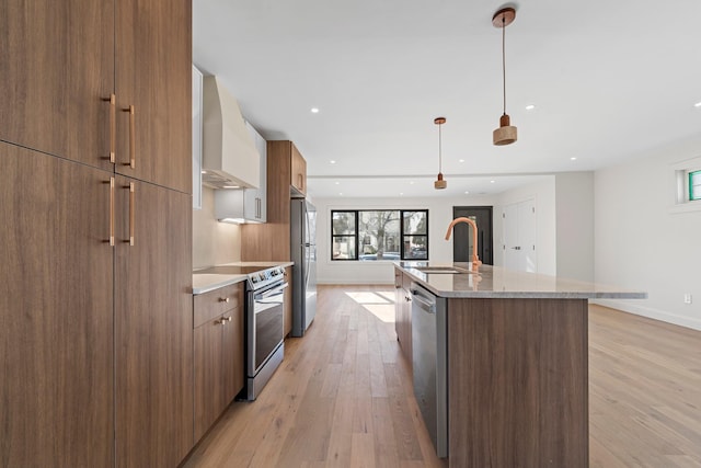 kitchen featuring appliances with stainless steel finishes, wall chimney range hood, a center island with sink, decorative light fixtures, and light hardwood / wood-style floors