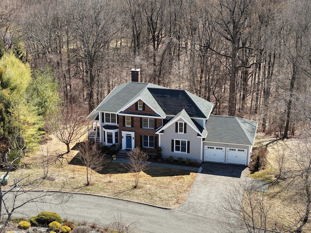 view of front of home featuring aphalt driveway, a wooded view, an attached garage, brick siding, and a chimney