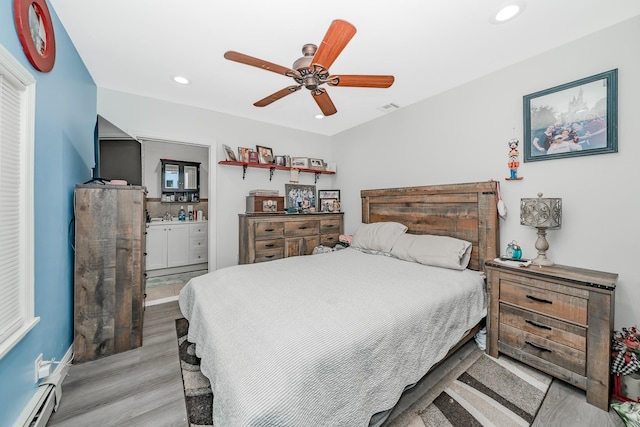 bedroom with ensuite bath, a baseboard radiator, ceiling fan, and light hardwood / wood-style flooring