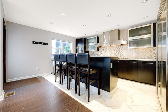 kitchen with a center island, backsplash, wall chimney exhaust hood, a kitchen bar, and dark brown cabinetry