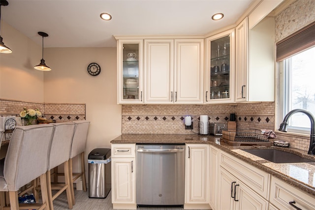 kitchen featuring sink, stainless steel dishwasher, cream cabinets, dark stone counters, and pendant lighting