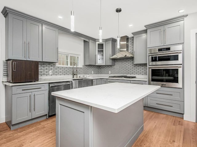 kitchen featuring wall chimney range hood, decorative backsplash, a kitchen island, gray cabinetry, and appliances with stainless steel finishes