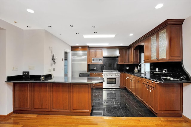 kitchen with wall chimney exhaust hood, built in appliances, tasteful backsplash, sink, and kitchen peninsula