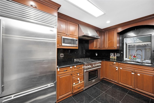 kitchen featuring wall chimney exhaust hood, dark stone counters, built in appliances, tasteful backsplash, and sink