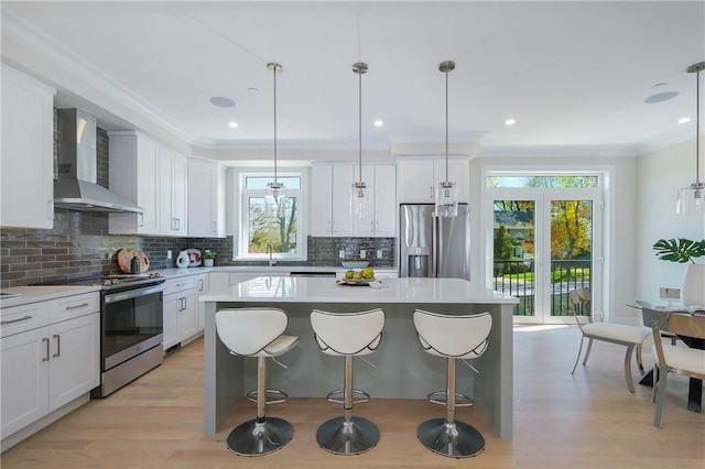 kitchen featuring appliances with stainless steel finishes, white cabinetry, wall chimney range hood, and decorative light fixtures