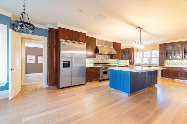 kitchen with extractor fan, hanging light fixtures, high quality appliances, a center island with sink, and ornamental molding