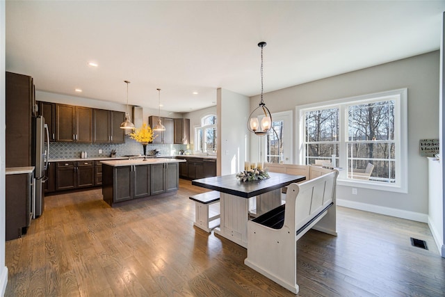 kitchen with hanging light fixtures, wall chimney range hood, a kitchen island, and stainless steel refrigerator