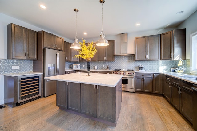 kitchen with wall chimney exhaust hood, decorative light fixtures, stainless steel appliances, wine cooler, and a center island with sink