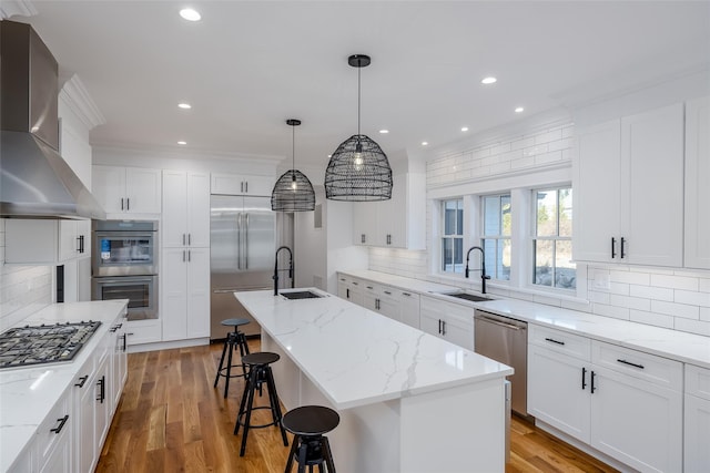 kitchen featuring stainless steel appliances, wall chimney exhaust hood, hanging light fixtures, and a kitchen island with sink