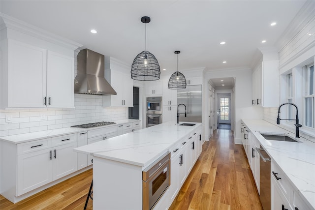 kitchen with pendant lighting, white cabinets, wall chimney exhaust hood, built in appliances, and a center island with sink