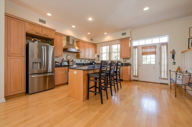 kitchen with stainless steel refrigerator with ice dispenser, a kitchen breakfast bar, wall chimney range hood, light hardwood / wood-style flooring, and a center island