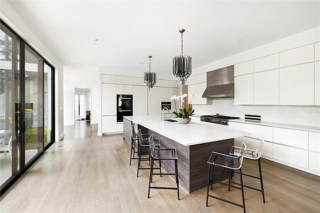 kitchen featuring decorative light fixtures, an island with sink, wall chimney range hood, light hardwood / wood-style floors, and a kitchen breakfast bar