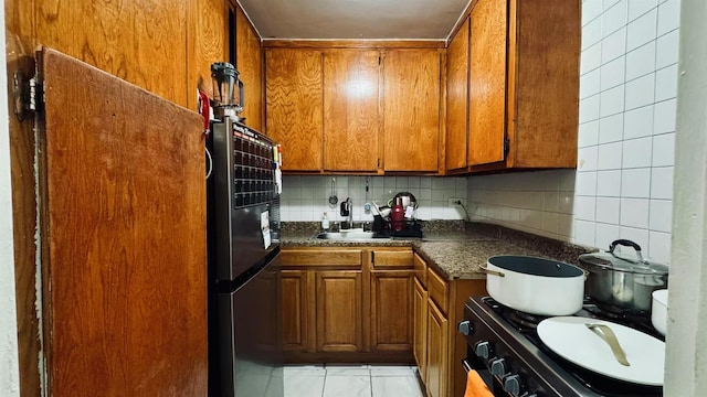 kitchen with black range oven, backsplash, light tile patterned flooring, stainless steel refrigerator, and sink