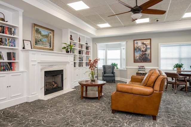 sitting room with a paneled ceiling, built in shelves, a fireplace, ornamental molding, and ceiling fan