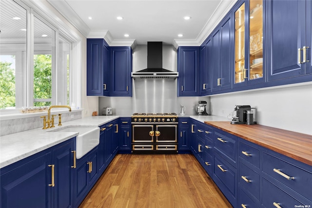 kitchen featuring blue cabinetry, butcher block counters, sink, and wall chimney range hood