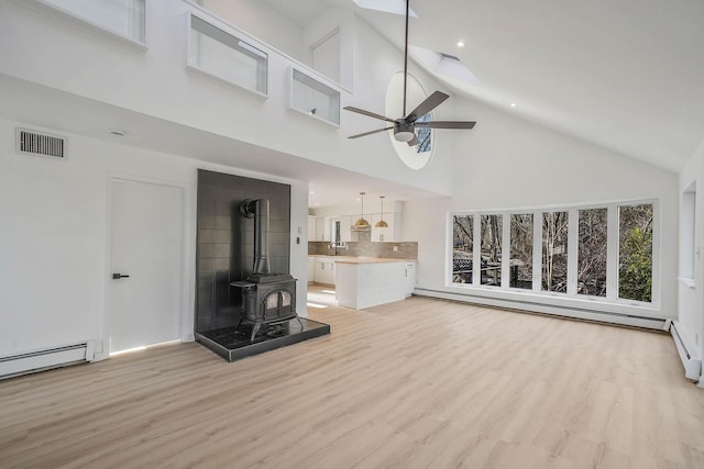 unfurnished living room featuring ceiling fan, a baseboard heating unit, a towering ceiling, light wood-type flooring, and a wood stove