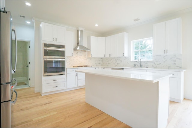 kitchen with light wood finished floors, wall chimney range hood, ornamental molding, stainless steel appliances, and a sink