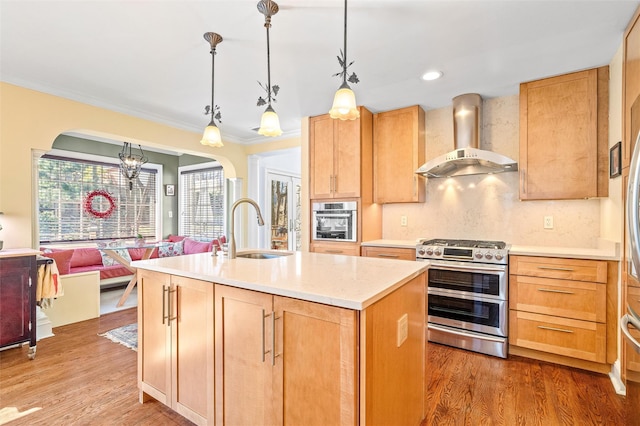 kitchen with arched walkways, light brown cabinetry, a sink, appliances with stainless steel finishes, and wall chimney exhaust hood