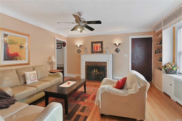 living room with a brick fireplace, ceiling fan, built in features, crown molding, and light hardwood / wood-style flooring