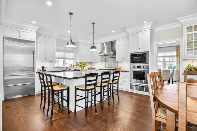 kitchen featuring a warming drawer, a breakfast bar, a sink, built in appliances, and wall chimney range hood