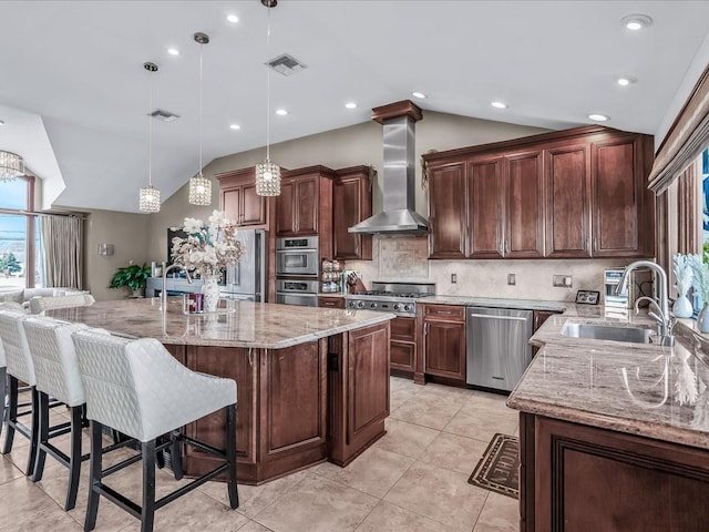 kitchen featuring pendant lighting, wall chimney exhaust hood, stainless steel appliances, a kitchen island with sink, and vaulted ceiling