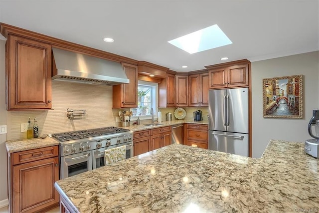 kitchen with light stone countertops, appliances with stainless steel finishes, wall chimney range hood, tasteful backsplash, and sink