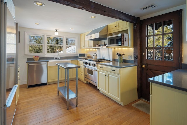 kitchen with wall chimney exhaust hood, sink, light hardwood / wood-style flooring, appliances with stainless steel finishes, and beamed ceiling