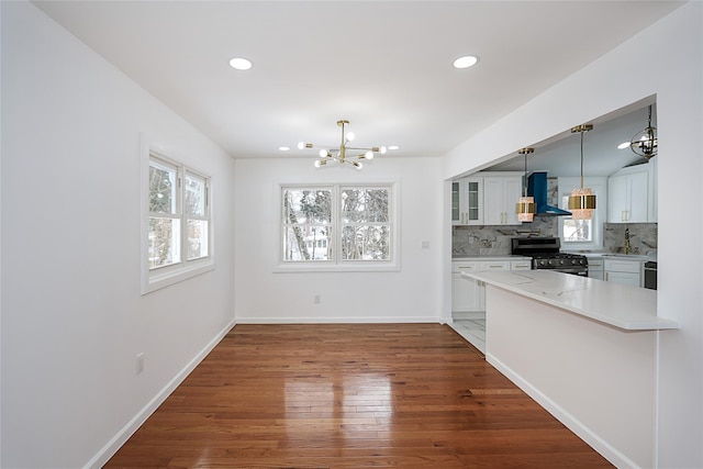 kitchen featuring kitchen peninsula, stainless steel range with gas cooktop, wall chimney exhaust hood, pendant lighting, and white cabinets