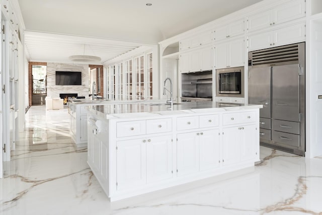 kitchen featuring stainless steel built in refrigerator, sink, white cabinets, a premium fireplace, and a center island with sink