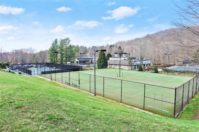 view of tennis court with a lawn, a forest view, and fence