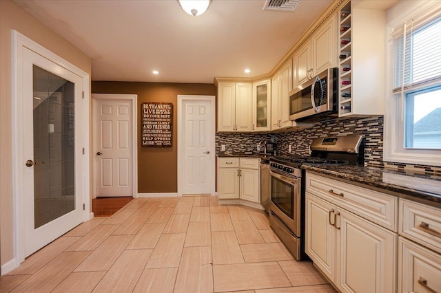 kitchen with backsplash, cream cabinetry, stainless steel appliances, and dark stone counters