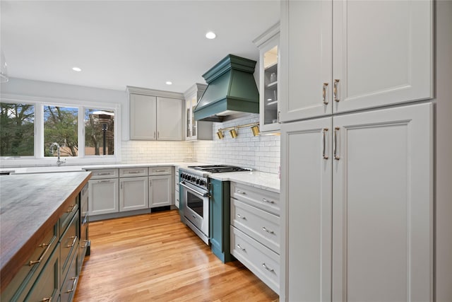 kitchen with custom exhaust hood, high end stainless steel range, backsplash, light stone countertops, and light hardwood / wood-style flooring