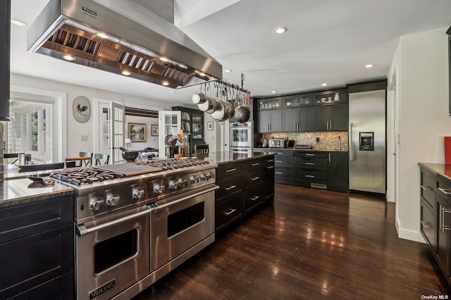 kitchen with appliances with stainless steel finishes, exhaust hood, dark hardwood / wood-style flooring, dark stone countertops, and backsplash