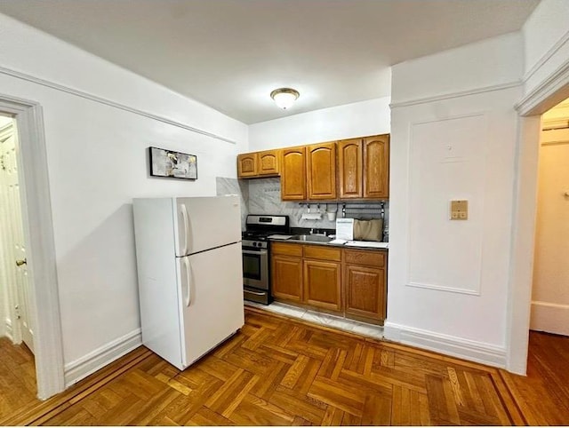 kitchen featuring backsplash, stainless steel range with gas cooktop, baseboards, freestanding refrigerator, and brown cabinetry