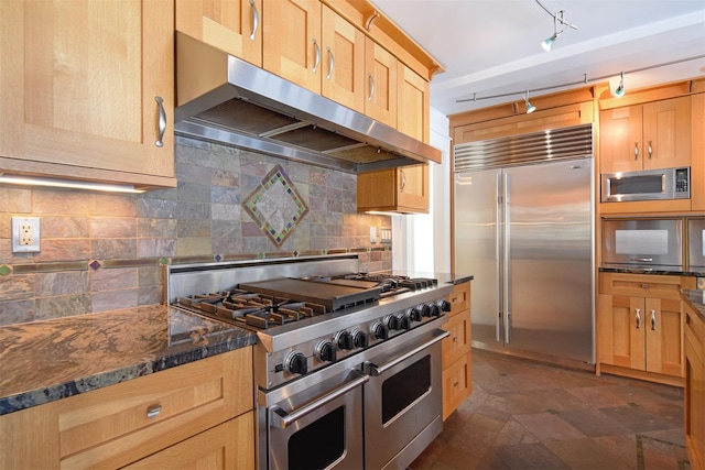 kitchen featuring rail lighting, built in appliances, dark stone countertops, range hood, and decorative backsplash