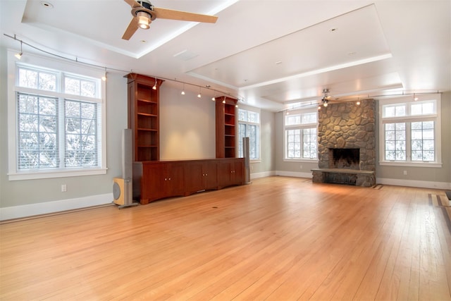 unfurnished living room with ceiling fan, a stone fireplace, a raised ceiling, and light hardwood / wood-style flooring