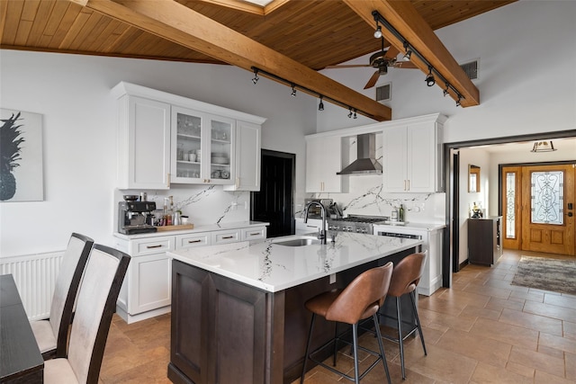 kitchen with white cabinetry, a kitchen island with sink, wall chimney range hood, track lighting, and lofted ceiling with beams