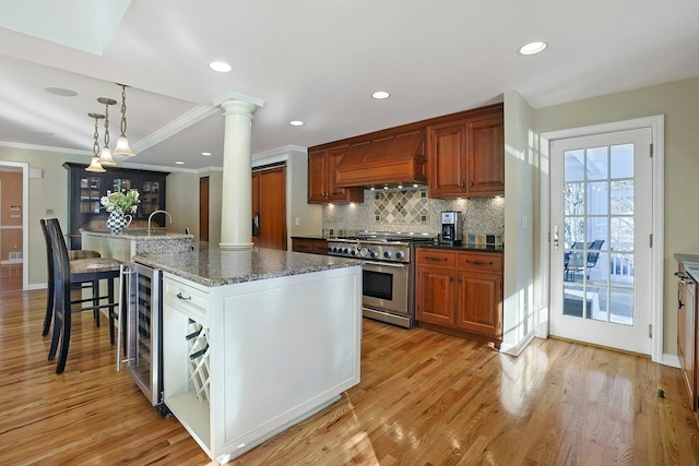 kitchen with premium range hood, ornate columns, hanging light fixtures, stainless steel stove, and a kitchen island with sink