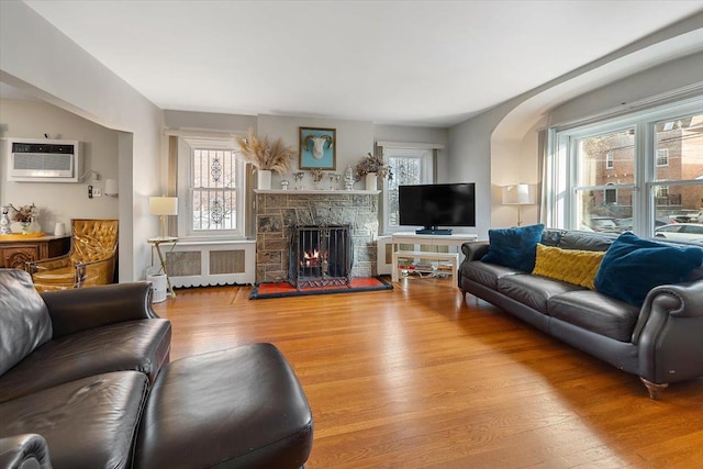 living room with a wall mounted air conditioner, a stone fireplace, light hardwood / wood-style floors, and radiator heating unit