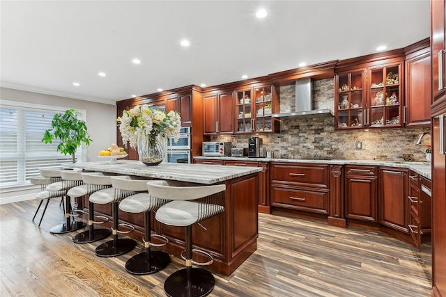 kitchen featuring wall chimney exhaust hood, a center island, double oven, light stone countertops, and backsplash