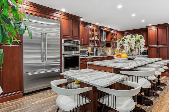 kitchen with a kitchen island, light stone counters, stainless steel appliances, light wood-type flooring, and wall chimney exhaust hood