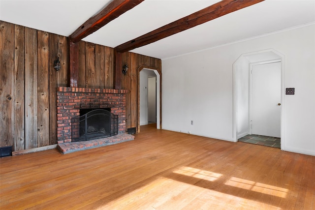 unfurnished living room featuring arched walkways, wood walls, a fireplace, wood finished floors, and beam ceiling