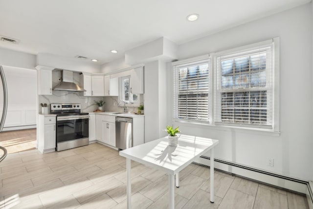 kitchen featuring appliances with stainless steel finishes, a baseboard radiator, wall chimney range hood, white cabinets, and sink