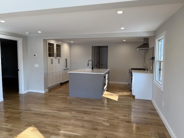 kitchen featuring light wood-style floors, light countertops, wall chimney exhaust hood, an island with sink, and gas range