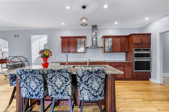 kitchen featuring double oven, backsplash, hanging light fixtures, light stone counters, and wall chimney exhaust hood