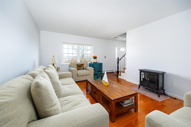 living area featuring baseboard heating, a wood stove, wood finished floors, baseboards, and stairs