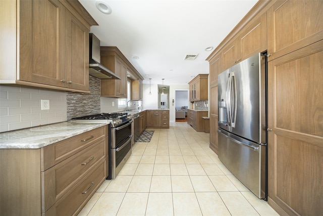 kitchen with appliances with stainless steel finishes, brown cabinetry, light tile patterned flooring, a sink, and wall chimney exhaust hood