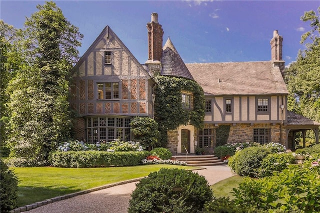 tudor home with stucco siding, stone siding, a chimney, and a front lawn