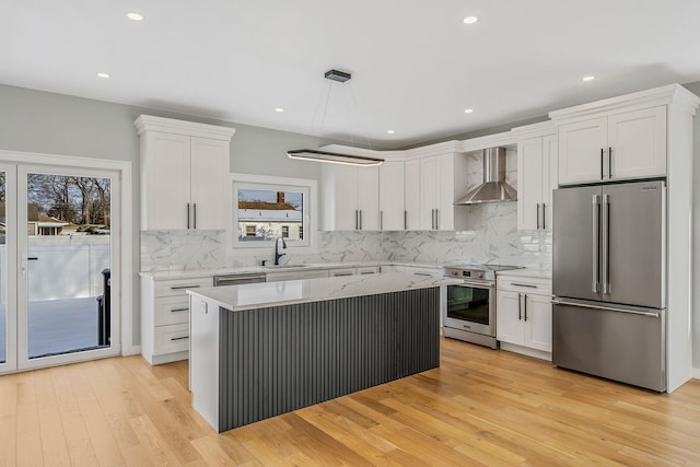 kitchen with appliances with stainless steel finishes, sink, white cabinets, hanging light fixtures, and wall chimney range hood